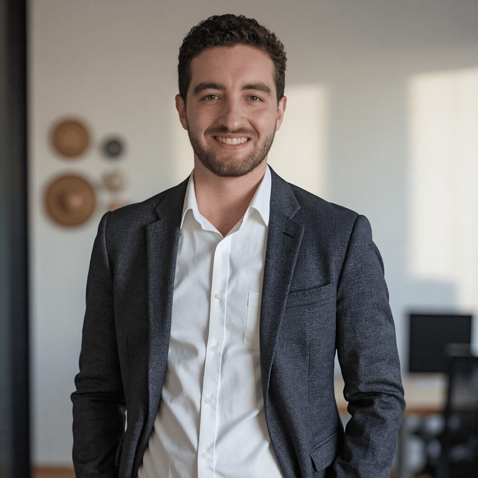 A casual professional photo of Hugo smiling in front of a modern office. He is sitting on a chair